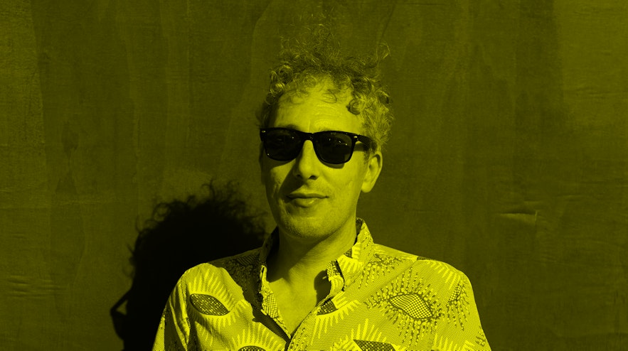 A person with short, grey, curly hair and light skin wearing sunglasses. They have a hint of a smile.