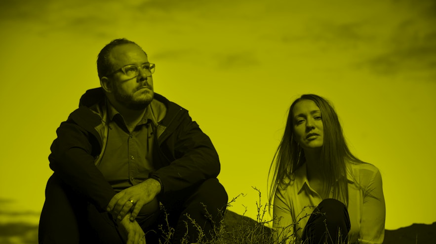 Two people sitting in a field at dusk. The person on the left has light skin, short hair, a moustache and glasses and gazes into the distance. The person on the right has long reddish hair and light skin and looks at you with their chin tilted up.