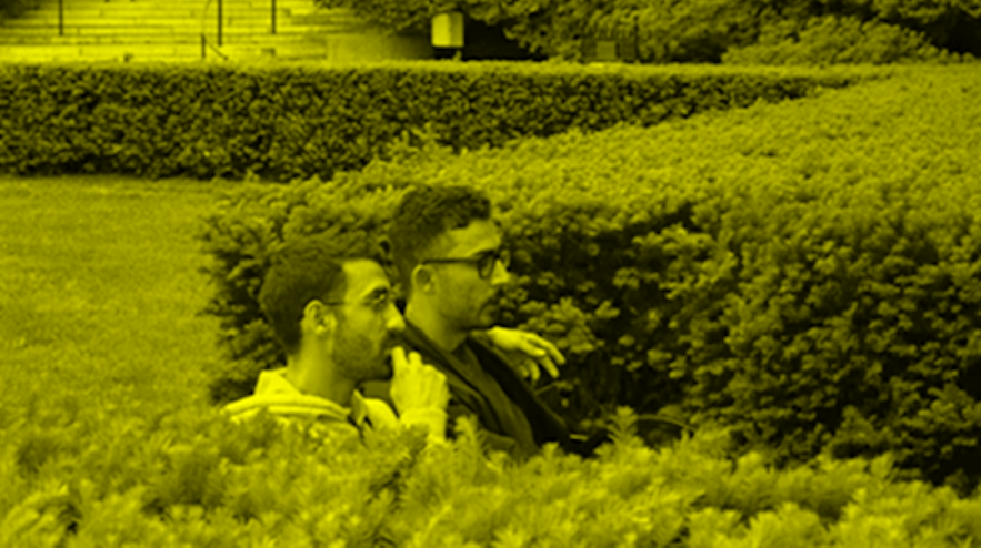 Two people seated, almost hidden, between two hedges in a park. Both have short black hair, light brown skin and glasses. The person on the right has their left arm around the person on the left.