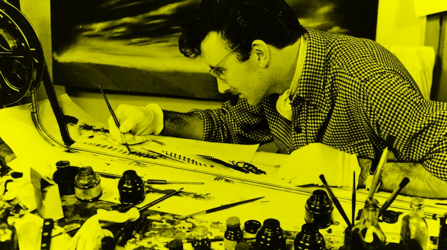 A person with dark, short hair and light skin painting on strips of celluloid film. In front of the person are many open bottles of ink.