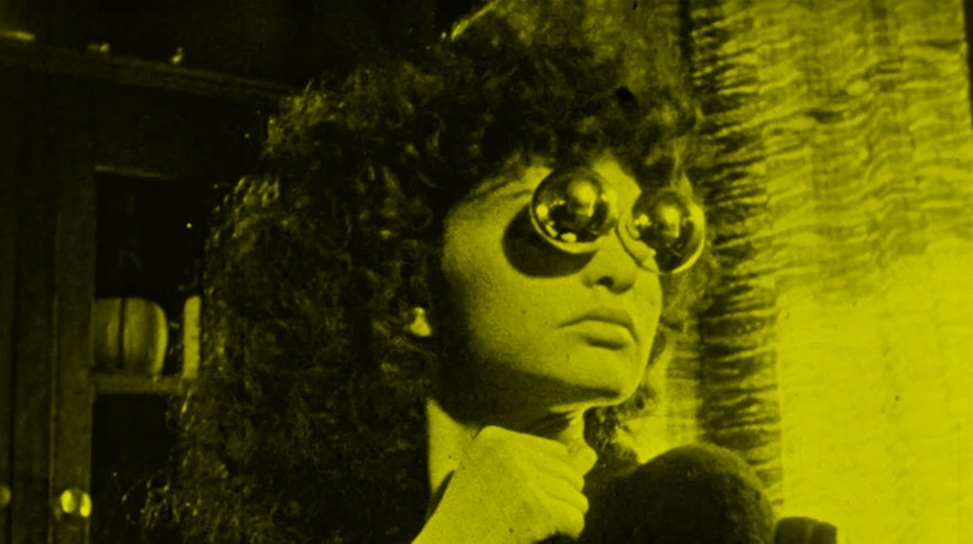 A person with voluminous dark, curly hair and light skin holds a knife in their right hand and is wearing spherical, silvery glasses.