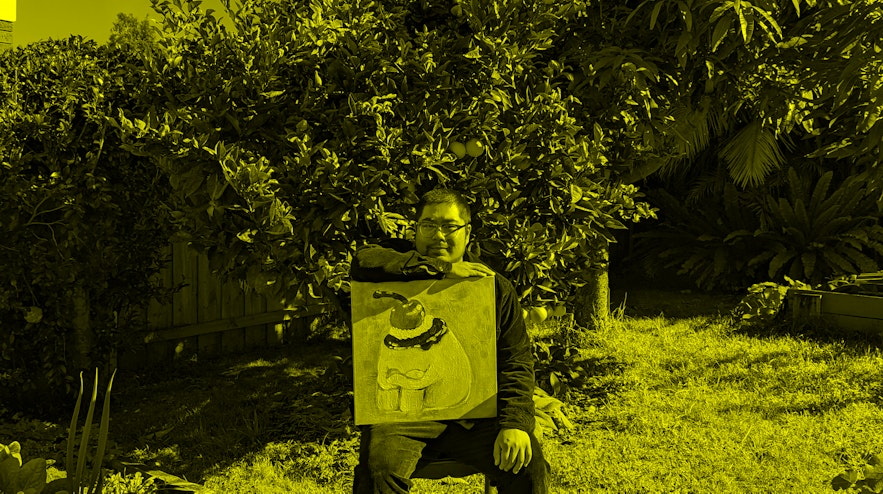 A person with short black hair and light skin sitting in a garden. They are resting their right hand on a canvas with a painting.