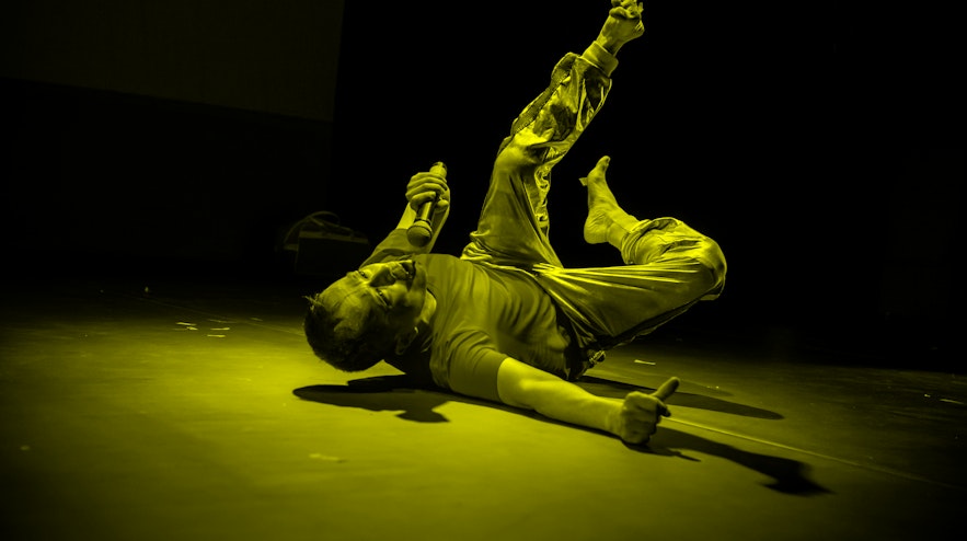 Person lying on their back on the floor with their legs in the air, one arm braced on the ground and the other holding a microphone to their mouth.