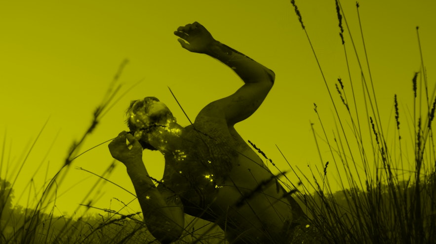 Person kneeling in tall grass outdoors, arms raised and leaning back to look at the sky.