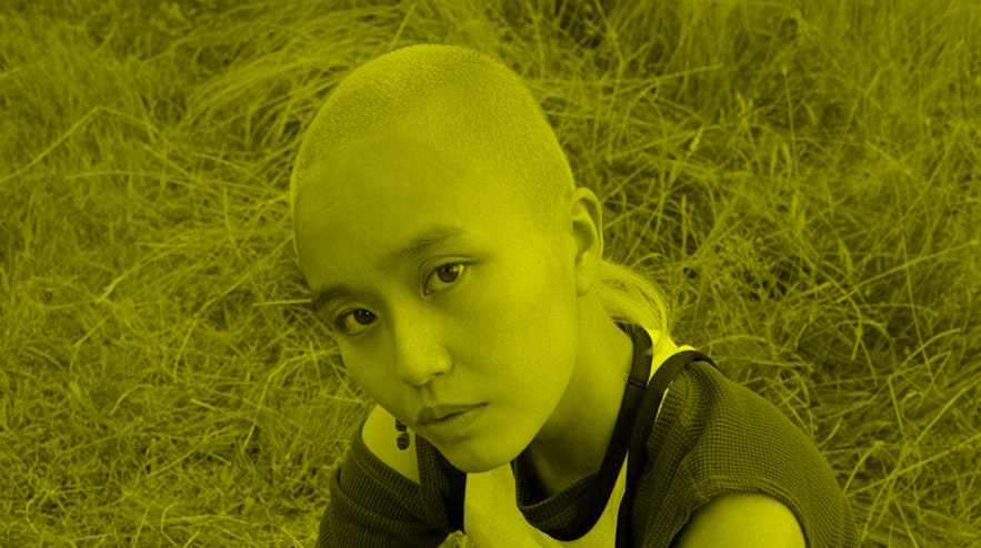 A person with light brown skin and a shaved head of green hair sits in a field looking up at you.