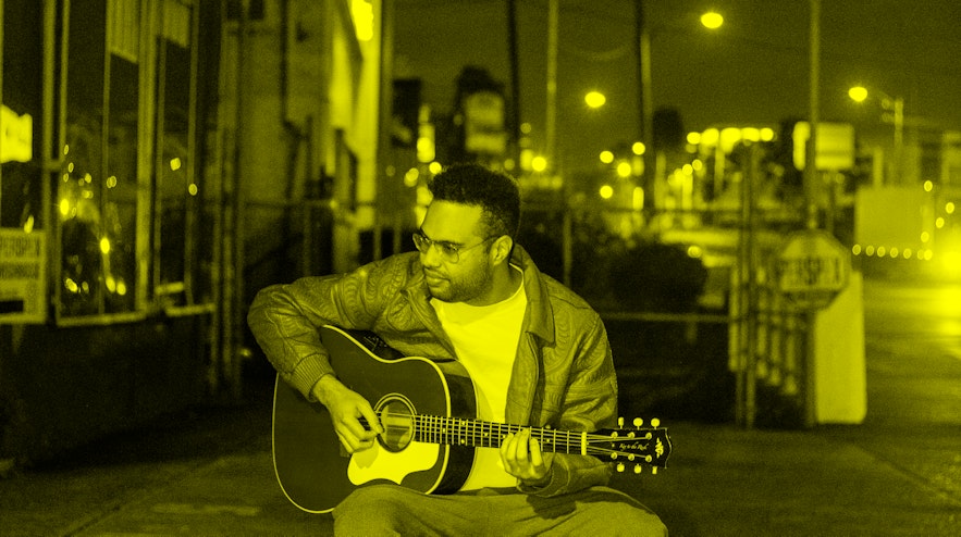 Person sitting on a red stool in a street playing an acoustic guitar