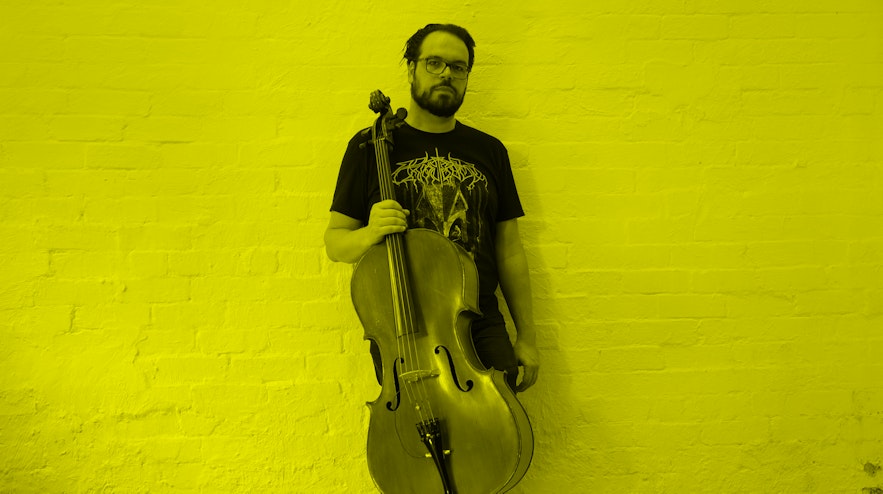 A person with light skin, dark short hair and beard holding a cello and leaning against the wall.